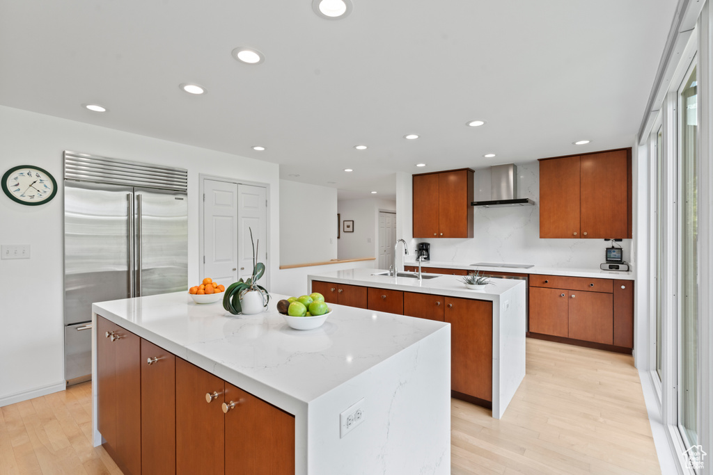 Kitchen featuring wall chimney exhaust hood, a sink, stainless steel built in refrigerator, light wood-style flooring, and an island with sink