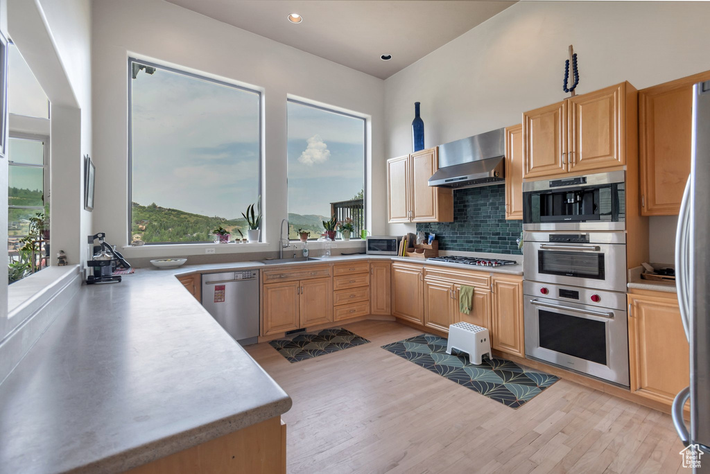 Kitchen with stainless steel appliances, a sink, light wood-style floors, wall chimney range hood, and light brown cabinets
