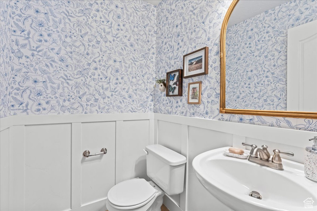 Half bathroom with a decorative wall, a sink, wallpapered walls, wainscoting, and toilet