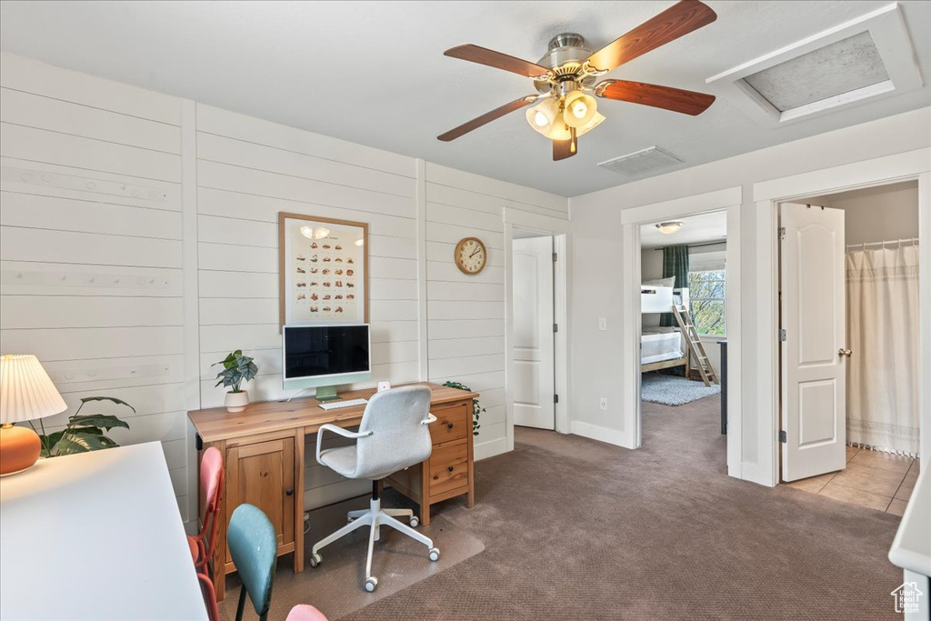 Carpeted home office featuring a ceiling fan, attic access, wooden walls, baseboards, and visible vents