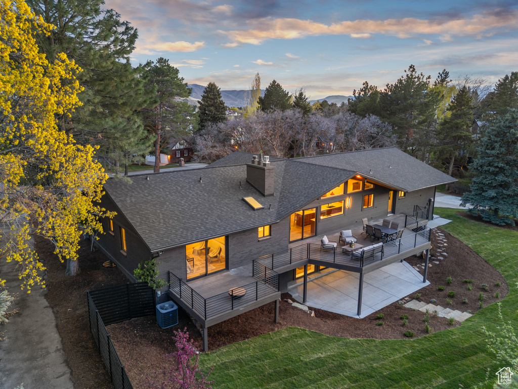 Back of property at dusk with central AC unit, a deck, a shingled roof, fence, and a yard