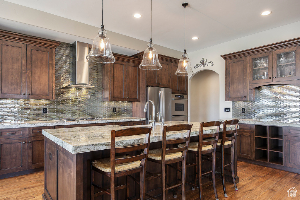 Kitchen with wall chimney exhaust hood, stainless steel appliances, tasteful backsplash, a breakfast bar, and light wood-type flooring