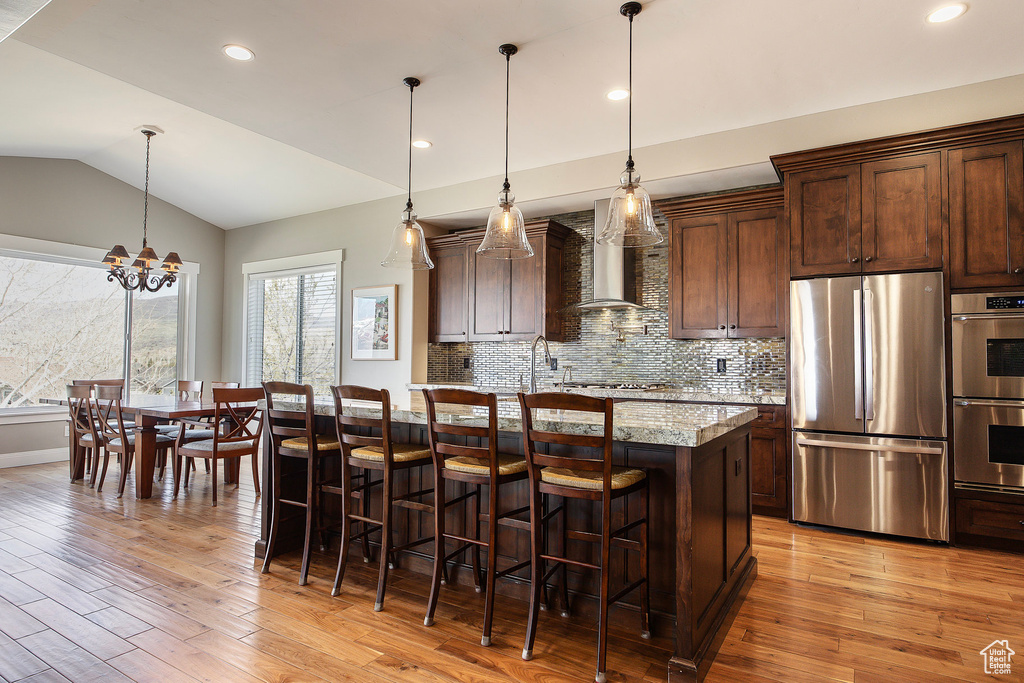 Kitchen featuring stainless steel appliances, wall chimney range hood, light wood-type flooring, a center island with sink, and lofted ceiling