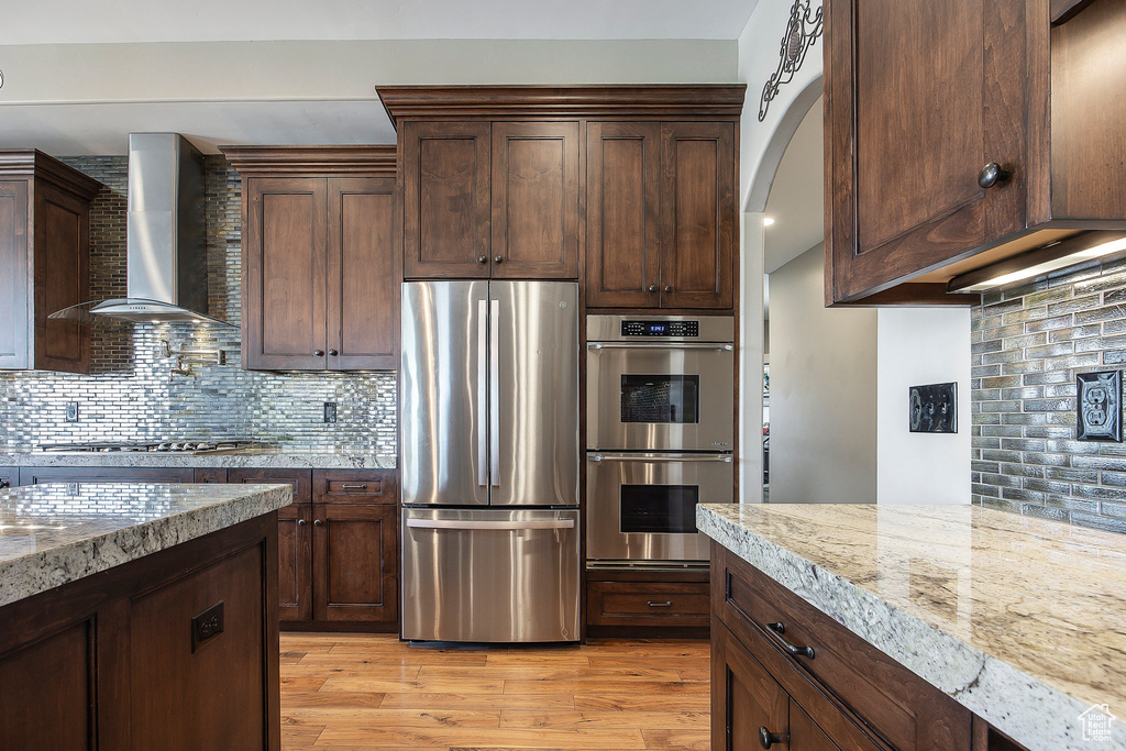Kitchen with appliances with stainless steel finishes, wall chimney exhaust hood, backsplash, light wood finished floors, and light stone countertops