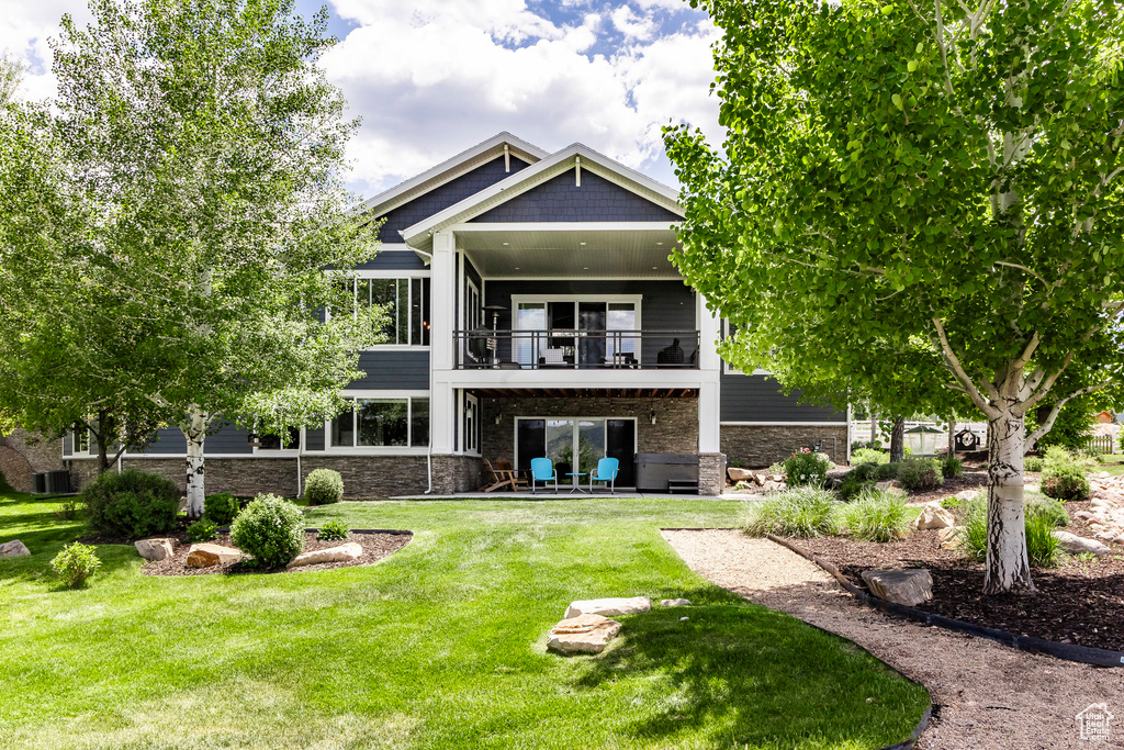 Rear view of house featuring a balcony, a lawn, a patio area, stone siding, and central air condition unit