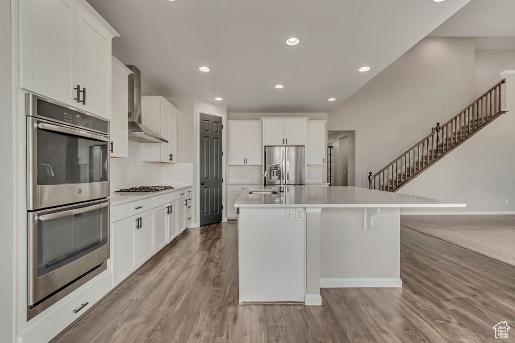 Kitchen with wall chimney exhaust hood, stainless steel appliances, an island with sink, light countertops, and dark wood finished floors