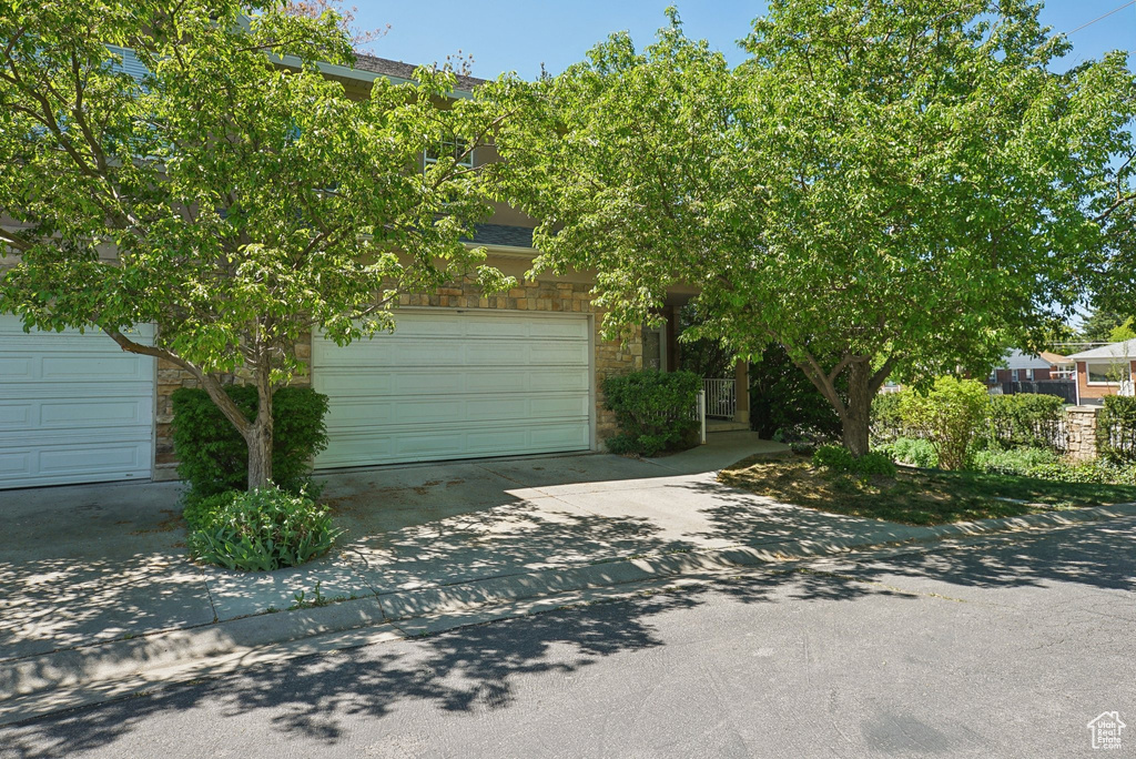 View of property hidden behind natural elements featuring an attached garage, a shingled roof, stone siding, and concrete driveway