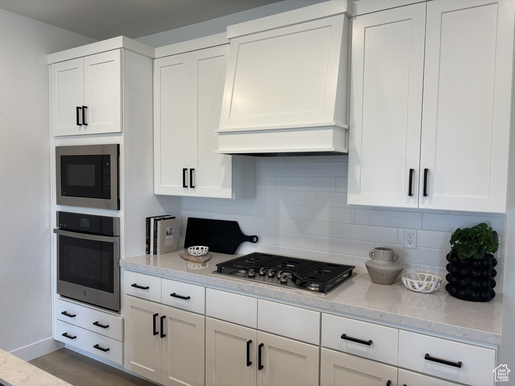 Kitchen with decorative backsplash, custom exhaust hood, white cabinetry, and appliances with stainless steel finishes