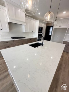 Kitchen featuring an island with sink, white cabinets, black stovetop, a sink, and dark wood-type flooring