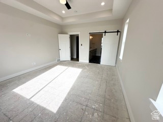 Unfurnished bedroom featuring a barn door, a ceiling fan, a tray ceiling, and baseboards