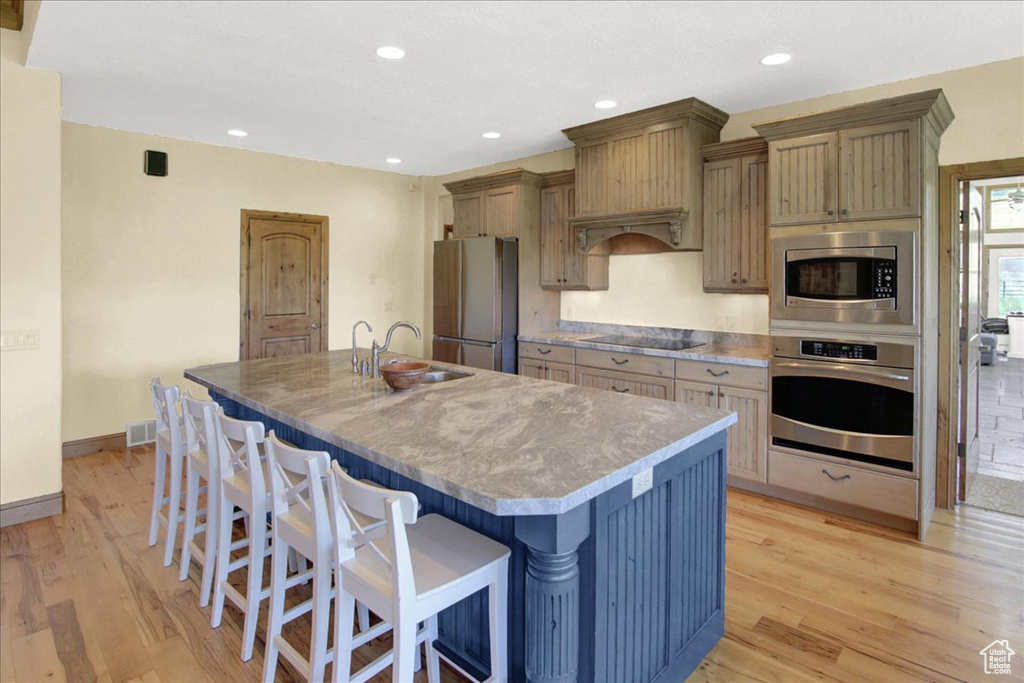Kitchen with a kitchen island with sink, light wood finished floors, a sink, stainless steel appliances, and a breakfast bar
