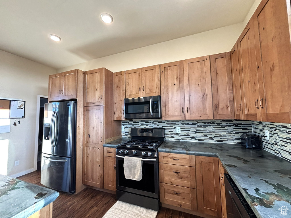 Kitchen with appliances with stainless steel finishes, dark countertops, dark wood-type flooring, brown cabinetry, and backsplash