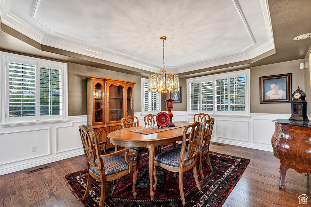 Dining space with dark wood finished floors, wainscoting, an inviting chandelier, crown molding, and a raised ceiling