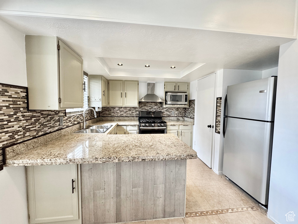 Kitchen with a sink, stainless steel appliances, a raised ceiling, wall chimney exhaust hood, and a peninsula