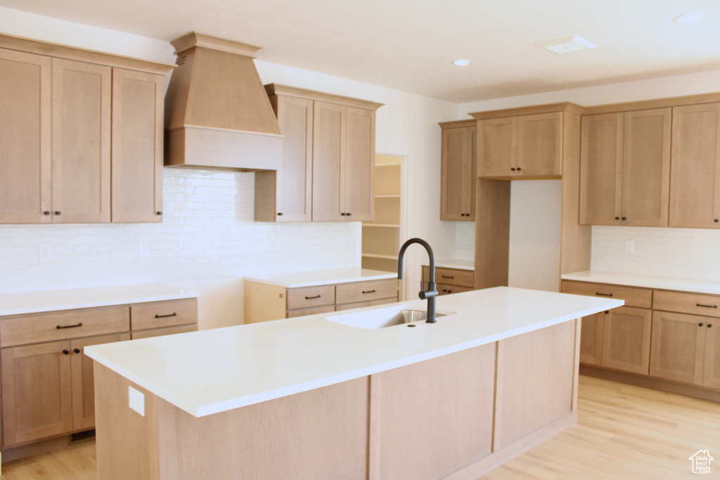 Kitchen with a sink, decorative backsplash, a center island with sink, light wood-style flooring, and premium range hood