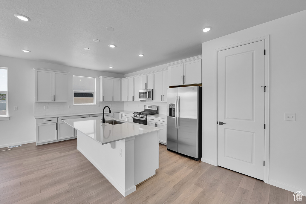 Kitchen featuring appliances with stainless steel finishes, white cabinets, light wood-style flooring, recessed lighting, and an island with sink
