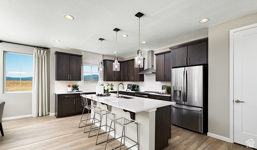 Kitchen with light wood finished floors, a sink, a breakfast bar, stainless steel refrigerator with ice dispenser, and wall chimney range hood