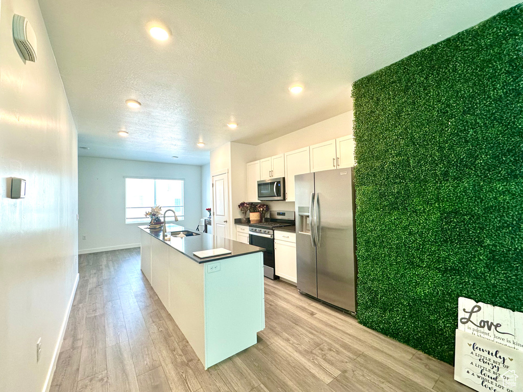 Kitchen featuring appliances with stainless steel finishes, white cabinets, light wood finished floors, dark countertops, and an island with sink