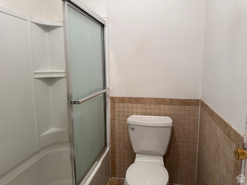 Full bath featuring tile walls, a wainscoted wall, bath / shower combo with glass door, and toilet