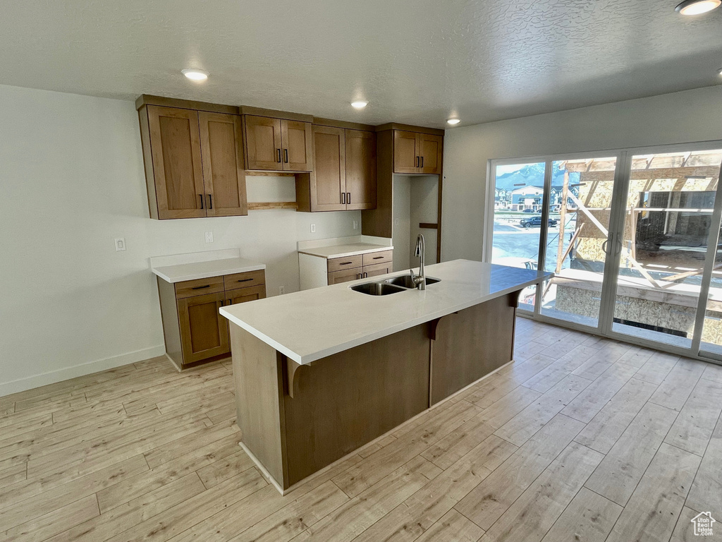 Kitchen featuring a sink, light wood finished floors, brown cabinetry, baseboards, and a center island with sink