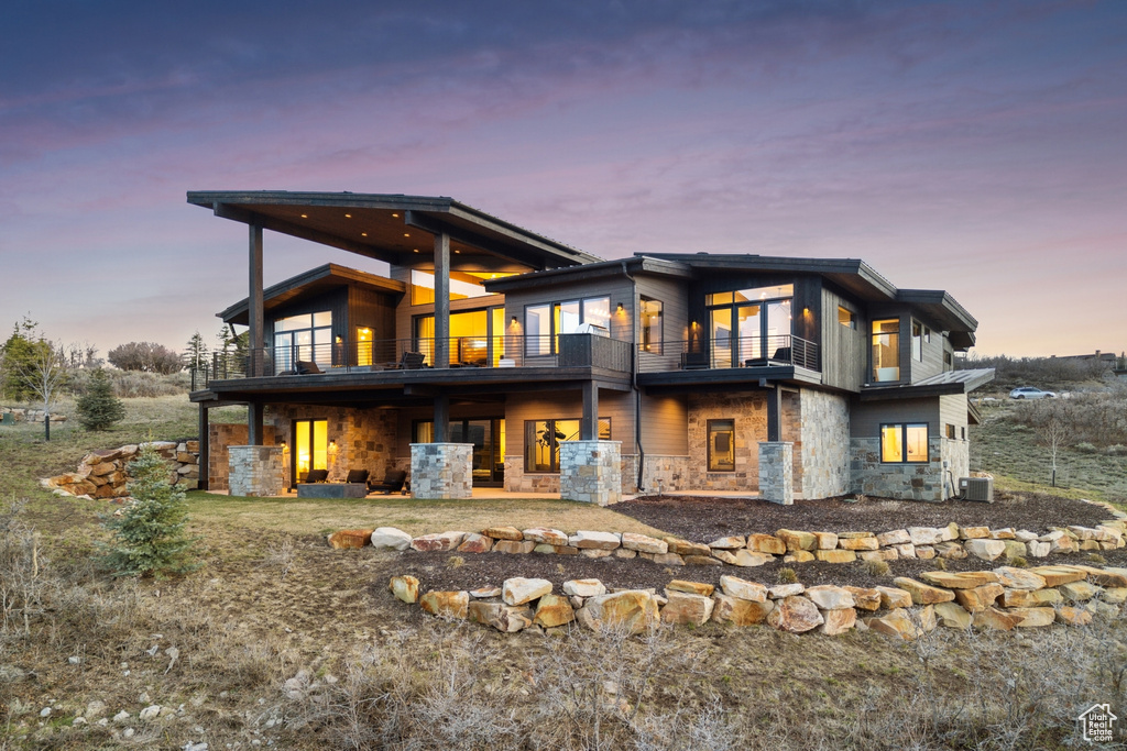 Back of property at dusk featuring a patio, central air condition unit, a balcony, and stone siding