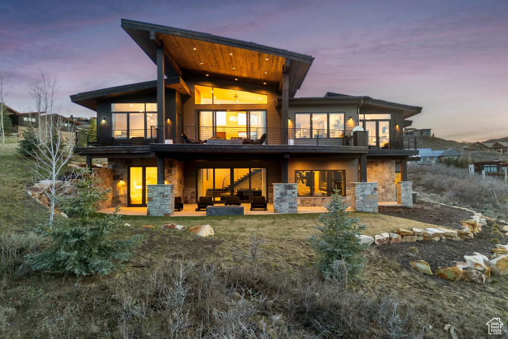 Back of house at dusk featuring a balcony, a patio, and stone siding