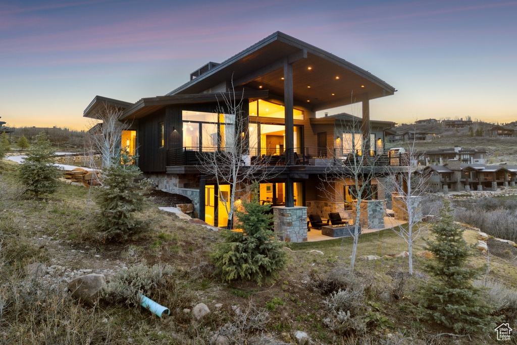 Back of house at dusk with a patio area, a balcony, and stone siding