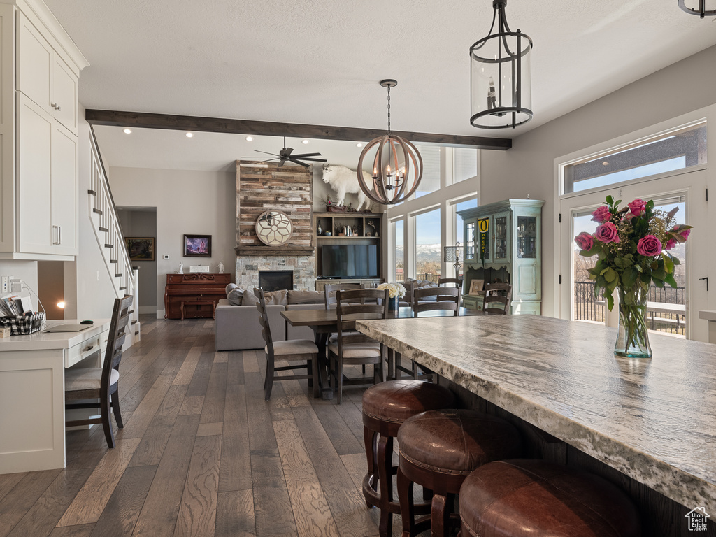 Dining space with dark wood-style flooring, stairway, beam ceiling, a stone fireplace, and ceiling fan with notable chandelier