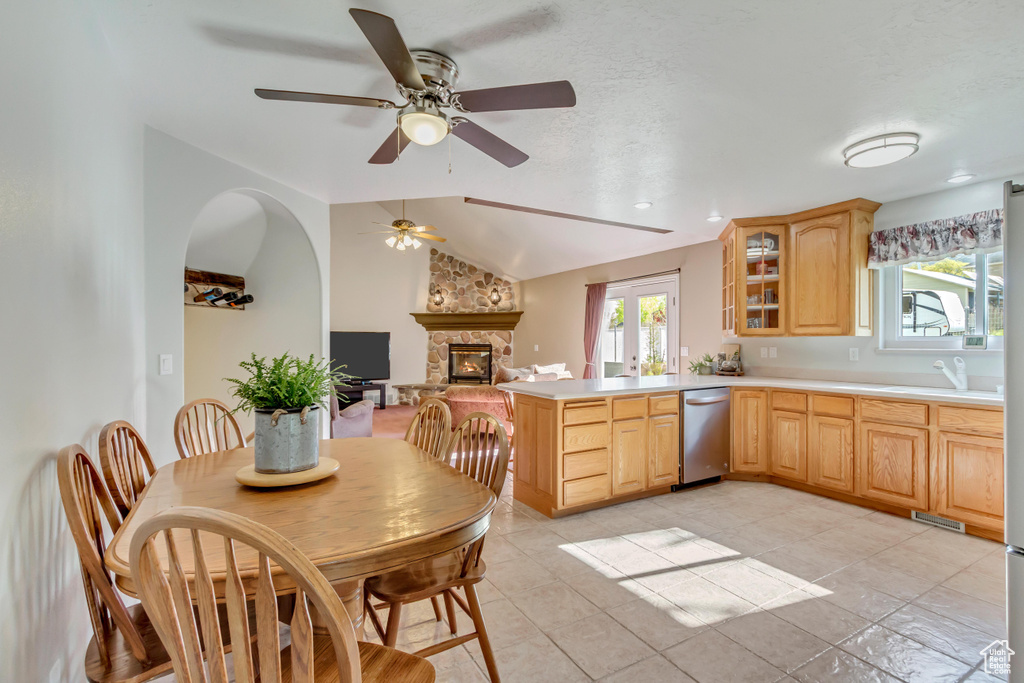 Kitchen featuring lofted ceiling, stainless steel dishwasher, light brown cabinetry, a ceiling fan, and a stone fireplace