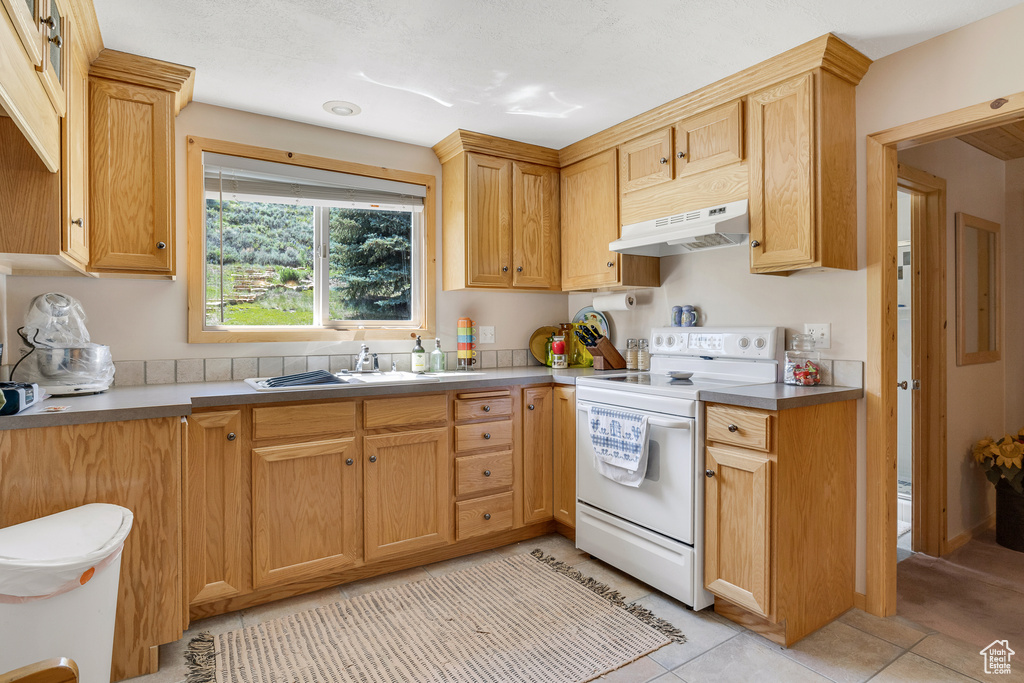 Kitchen with white electric range oven, light tile patterned flooring, under cabinet range hood, and a sink
