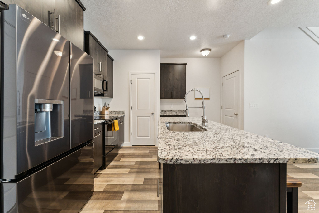 Kitchen with dark brown cabinetry, wood finished floors, black appliances, a sink, and a center island with sink