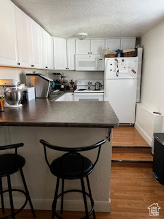 Kitchen with a textured ceiling, a breakfast bar, dark countertops, white appliances, and a peninsula