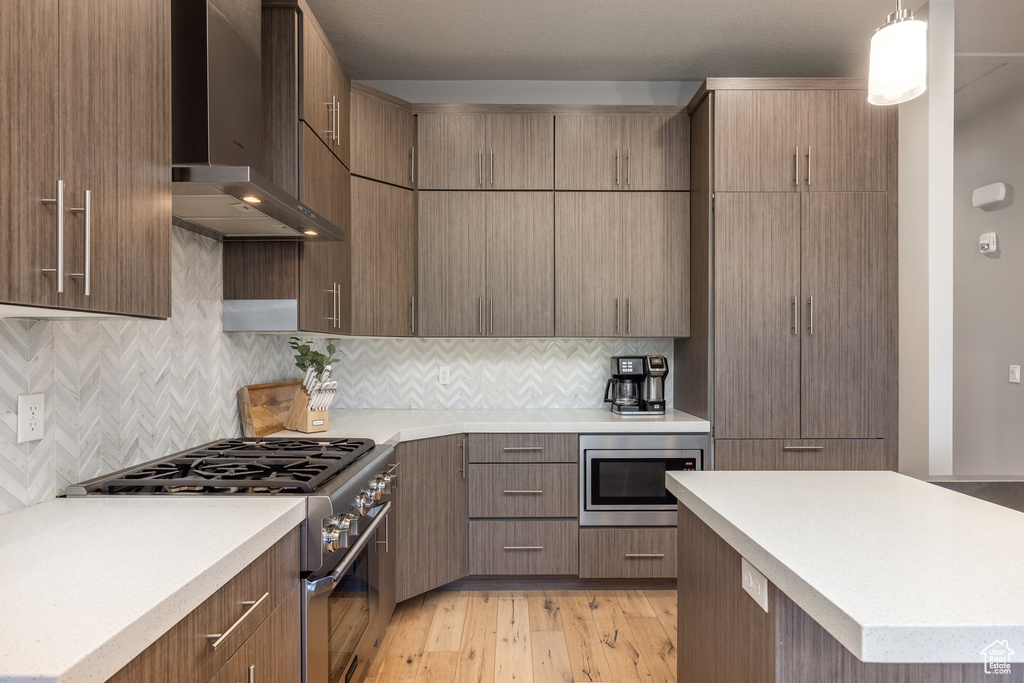 Kitchen with tasteful backsplash, stainless steel appliances, light countertops, light wood-type flooring, and wall chimney exhaust hood