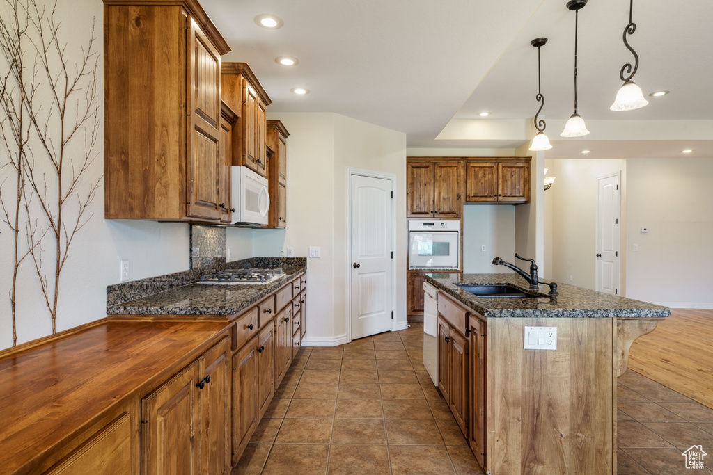 Kitchen featuring white appliances, brown cabinetry, a sink, baseboards, and a center island with sink
