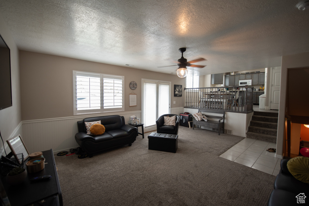 Living area with a ceiling fan, a textured ceiling, wainscoting, light carpet, and stairs