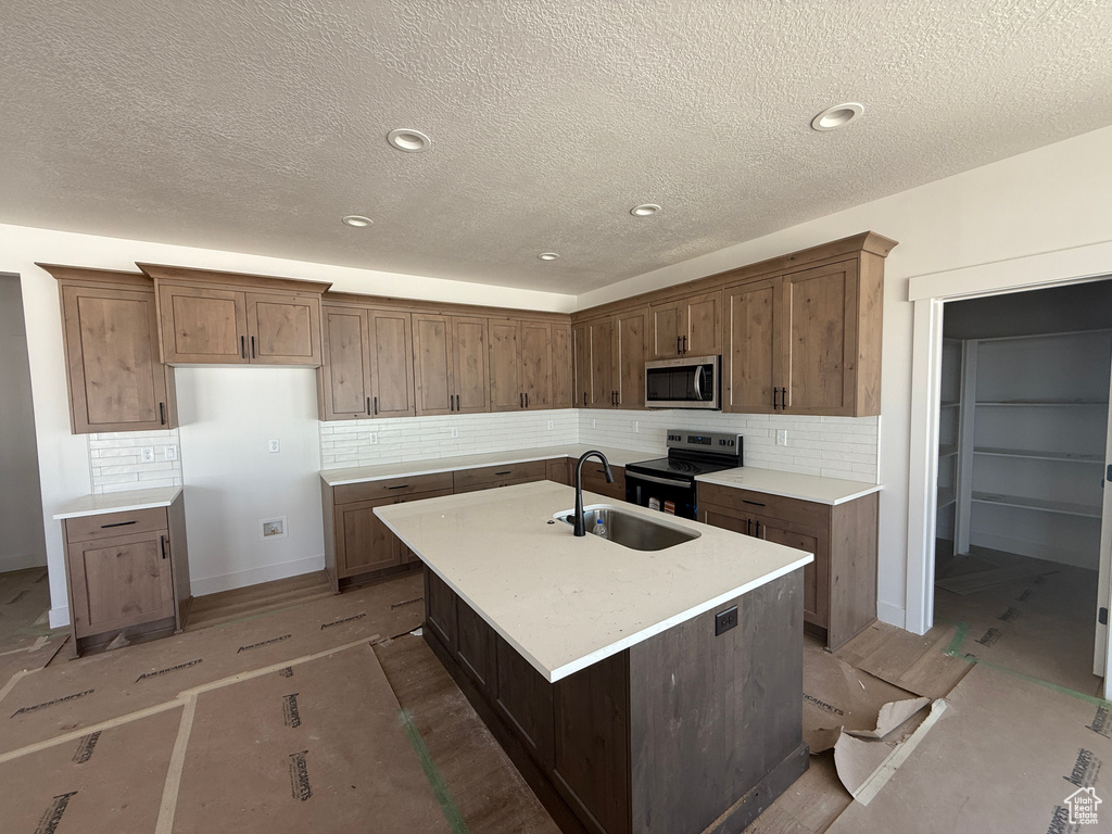 Kitchen featuring appliances with stainless steel finishes, backsplash, an island with sink, light countertops, and a textured ceiling