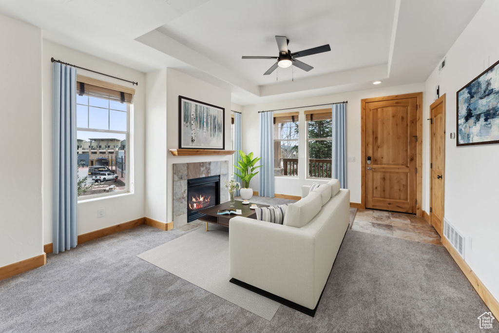 Carpeted living area featuring a tiled fireplace, a raised ceiling, ceiling fan, and a healthy amount of sunlight