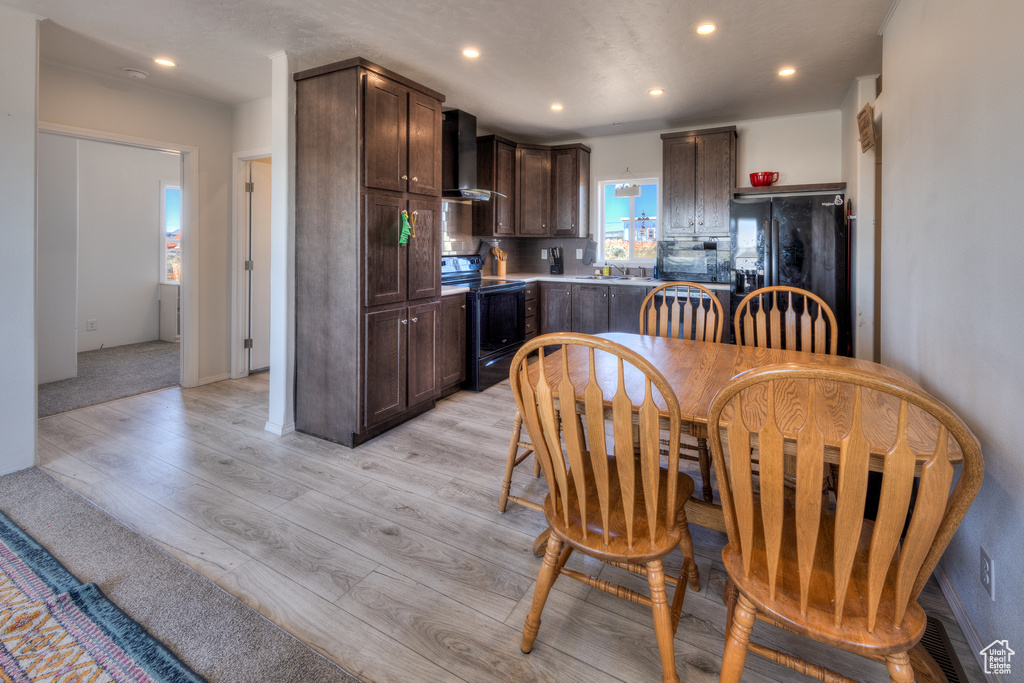 Kitchen with black appliances, light wood-style floors, dark brown cabinetry, wall chimney exhaust hood, and recessed lighting