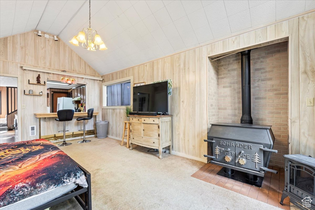 Carpeted bedroom with a wood stove, wood walls, freestanding refrigerator, a notable chandelier, and vaulted ceiling