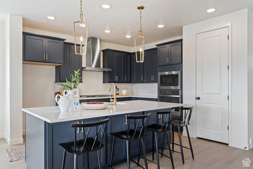 Kitchen featuring a breakfast bar area, backsplash, light wood finished floors, stainless steel appliances, and wall chimney exhaust hood