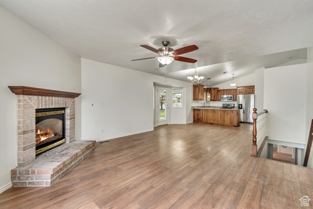 Unfurnished living room featuring a brick fireplace, ceiling fan with notable chandelier, baseboards, wood finished floors, and vaulted ceiling