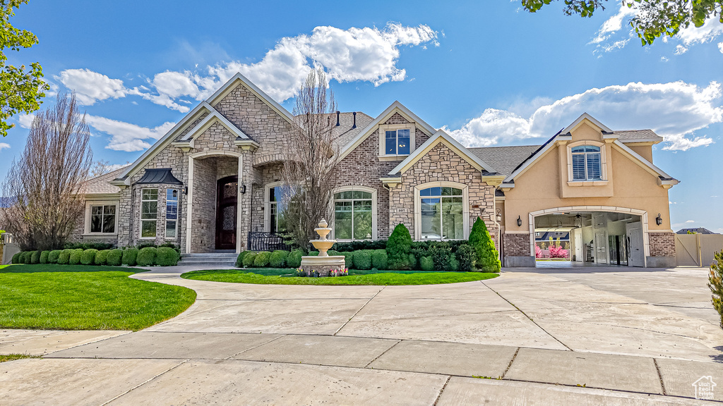 French country inspired facade with a front yard, curved driveway, an attached garage, stone siding, and stucco siding