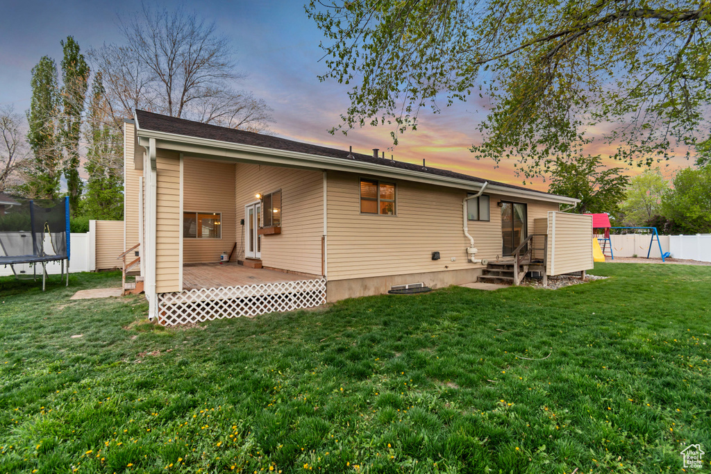 Back of house with fence, a trampoline, a playground, a wooden deck, and a lawn