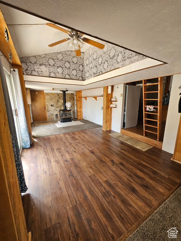 Unfurnished living room featuring a wood stove, vaulted ceiling, hardwood / wood-style floors, ceiling fan, and a textured ceiling