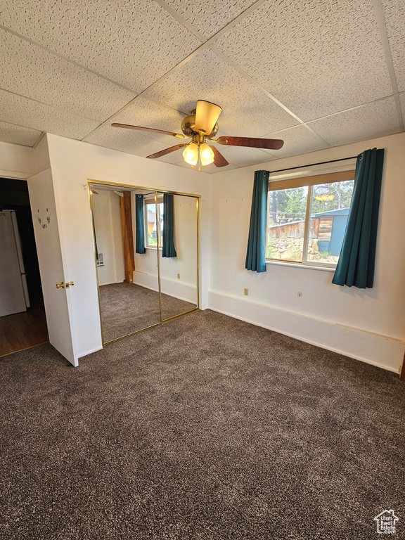 Unfurnished bedroom featuring freestanding refrigerator, a drop ceiling, dark carpet, a closet, and ceiling fan
