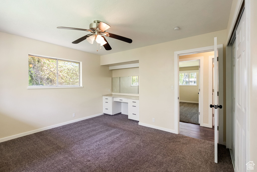 Unfurnished bedroom with ceiling fan, built in study area, dark colored carpet, baseboards, and a closet