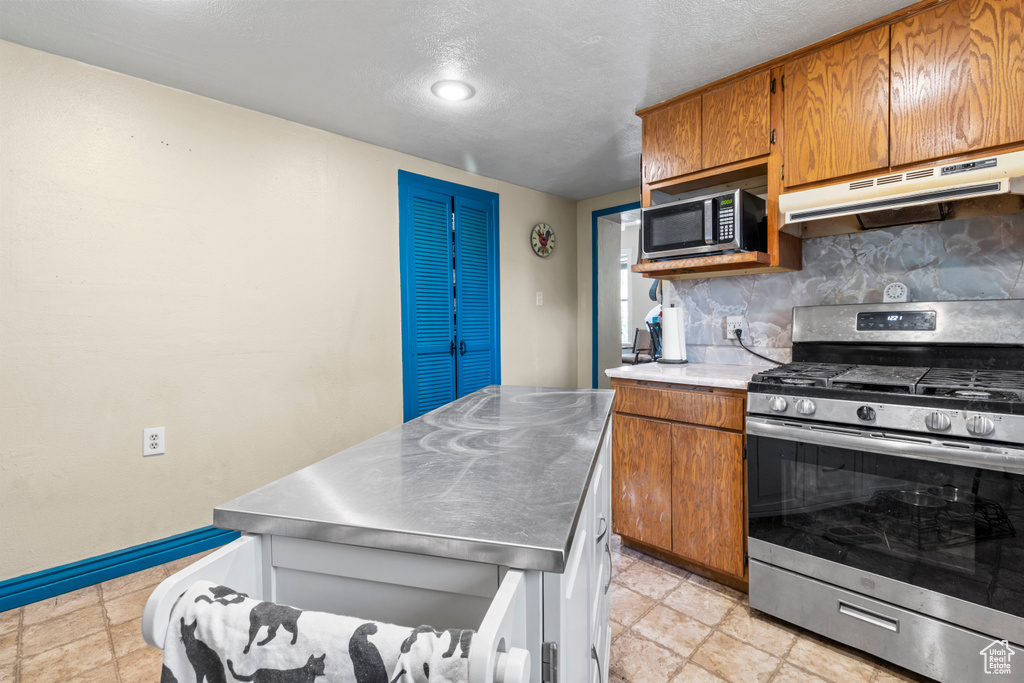 Kitchen featuring stainless steel countertops, appliances with stainless steel finishes, under cabinet range hood, brown cabinets, and backsplash