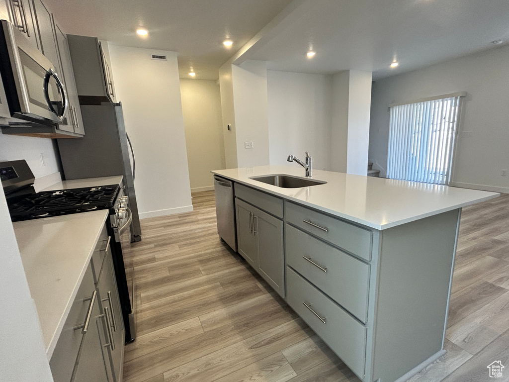 Kitchen featuring a sink, gray cabinetry, light wood-style floors, stainless steel appliances, and an island with sink