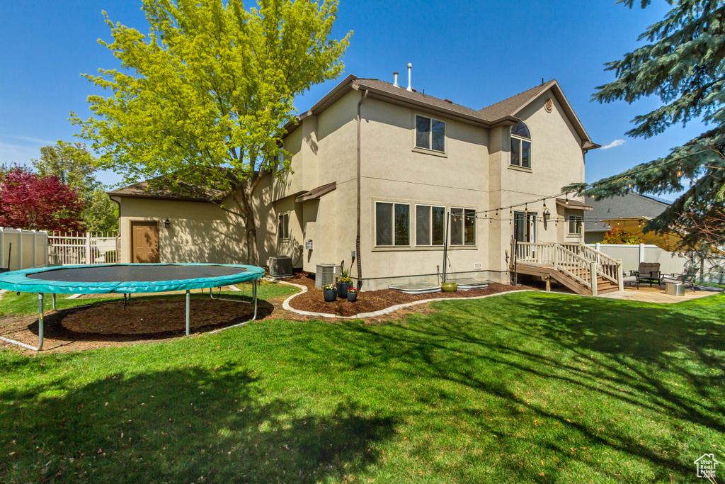 Rear view of property with fence, a trampoline, a lawn, stucco siding, and central AC unit