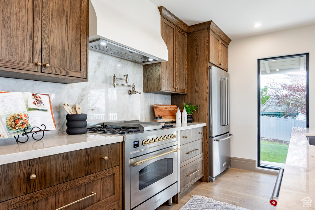 Kitchen with wall chimney range hood, light stone counters, light wood-style floors, tasteful backsplash, and premium appliances